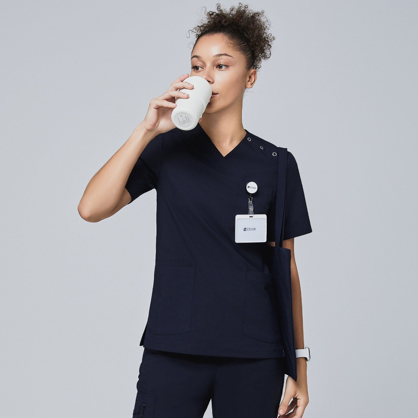Image of Model in navy V-neck scrub top with shoulder buttons, drinking from a cup and wearing a Zenir badge,Eco Navy